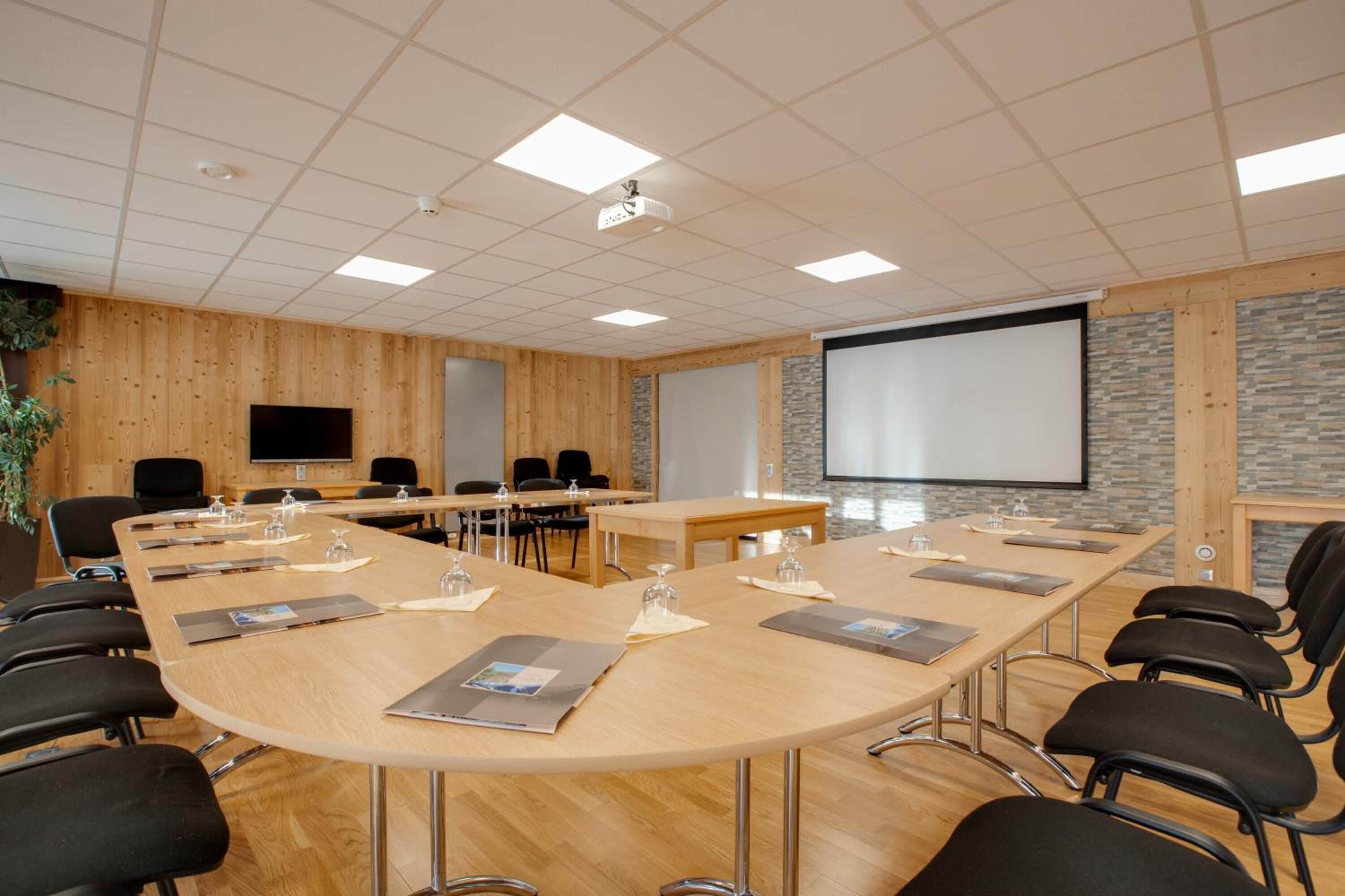 Una sala de conferències amb una taula llarga, cadires negres, una pantalla de projector i una paret de pedra decorativa.