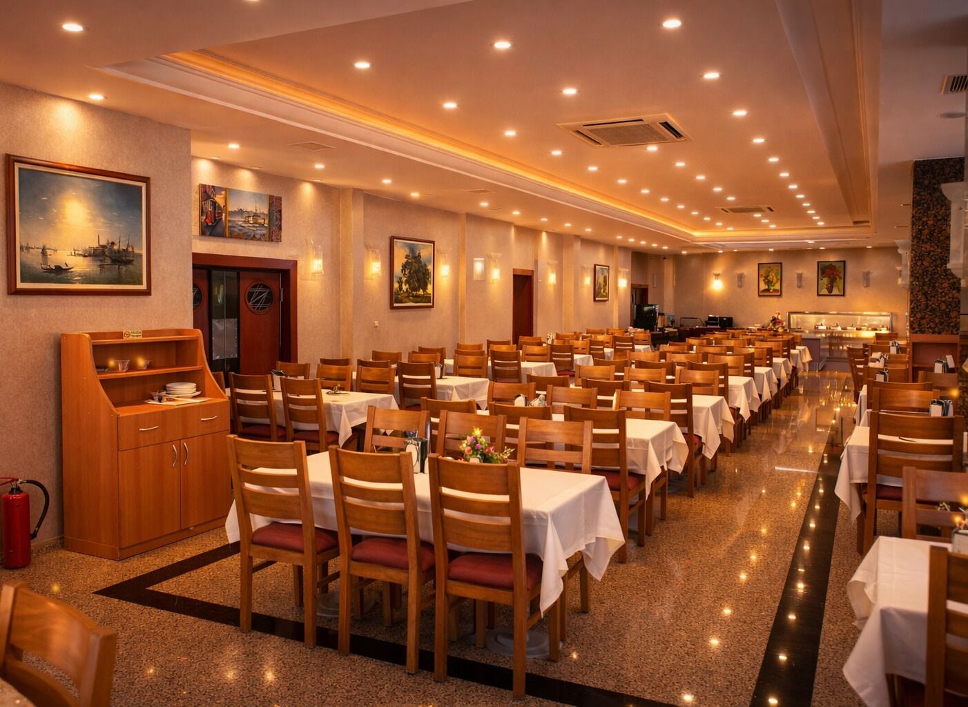 A spacious dining hall with rows of tables and chairs, a buffet area, and artwork on the walls.