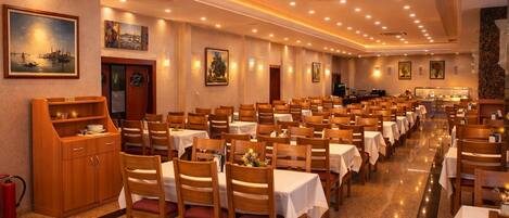 A spacious dining hall with rows of tables and chairs, a buffet area, and artwork on the walls.