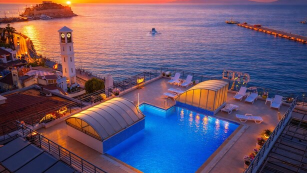 A rooftop pool with a clock tower, lounge chairs, and a view of the sea at sunset. - Kayhanbey Otel (Kuşadası)