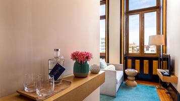 Ein Hotelzimmer mit einem Holztisch, einer Spirituosenflasche, Gläsern, einer Blumenvase, einer Couch, einer Lampe und Blick auf Gebäude durch große Fenster.