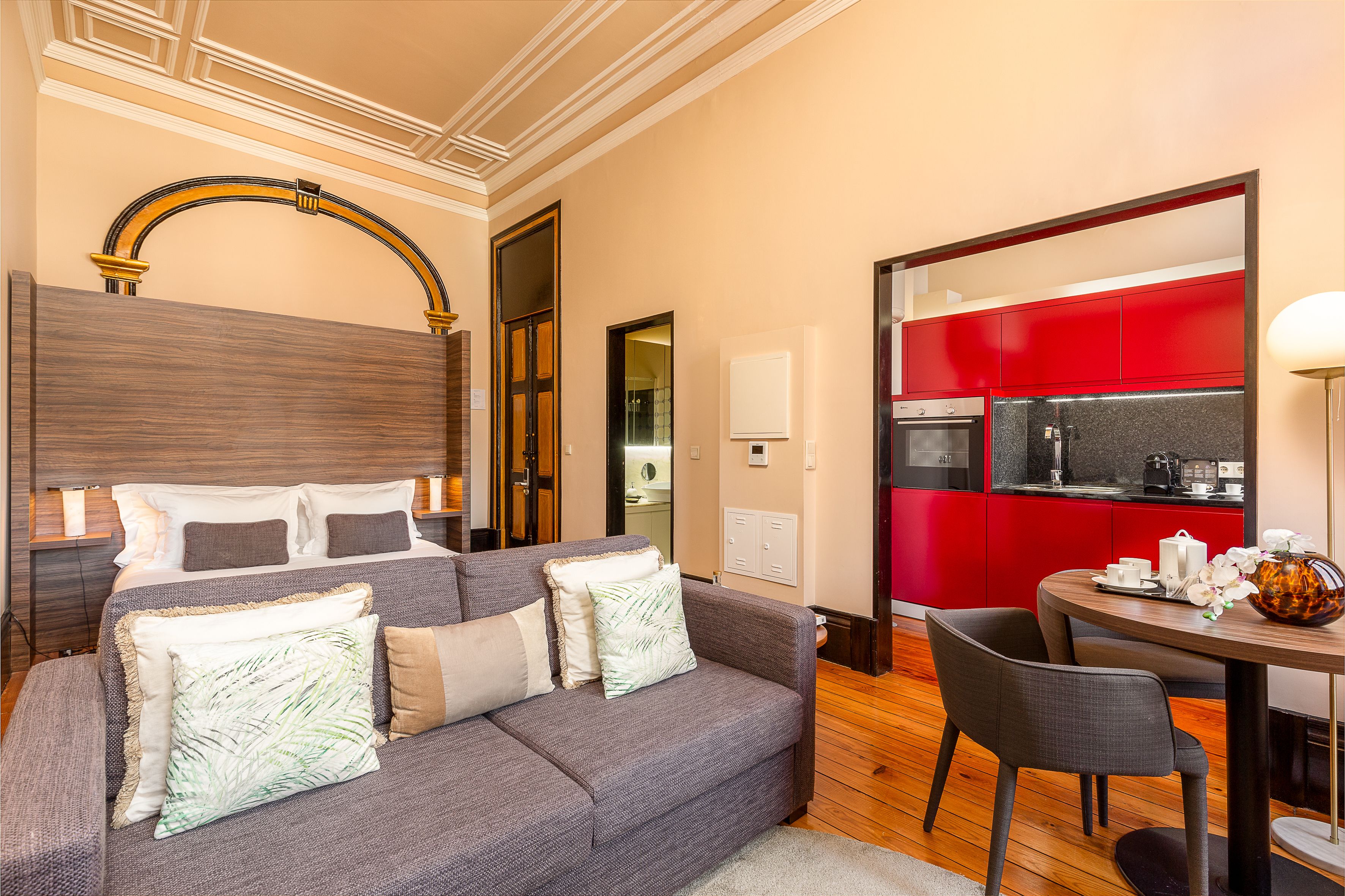 Une chambre d’hôtel moderne avec un canapé gris, une tête de lit en bois, un coin cuisine rouge et une table à manger avec des chaises.