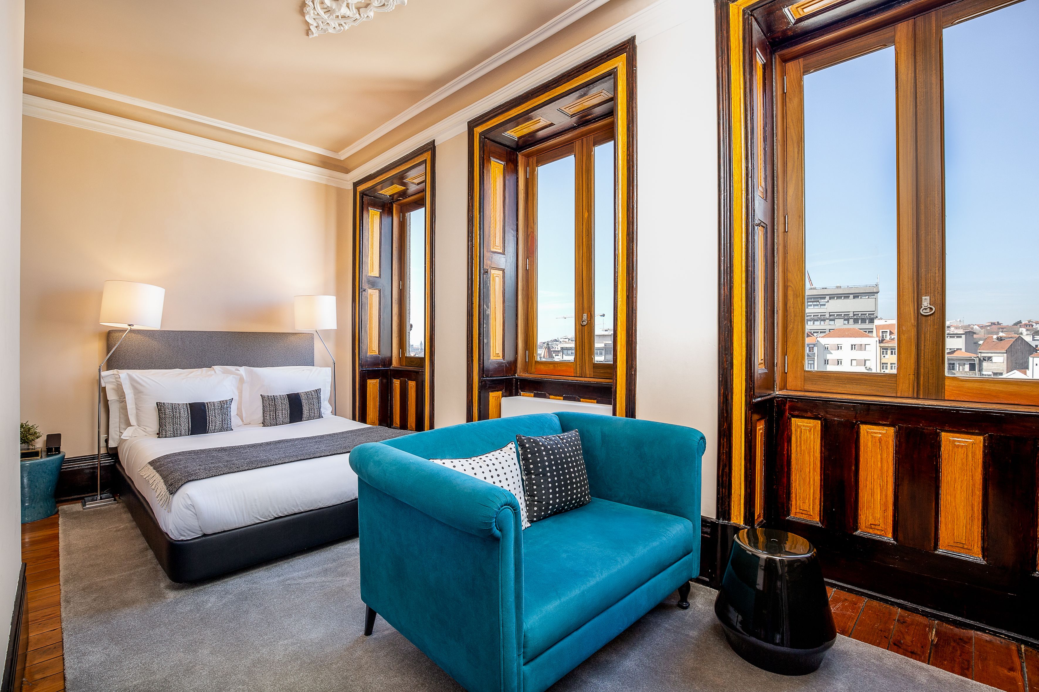 Una habitación de hotel con una cama, un sofá color turquesa y ventanas amplias con vista a la ciudad.