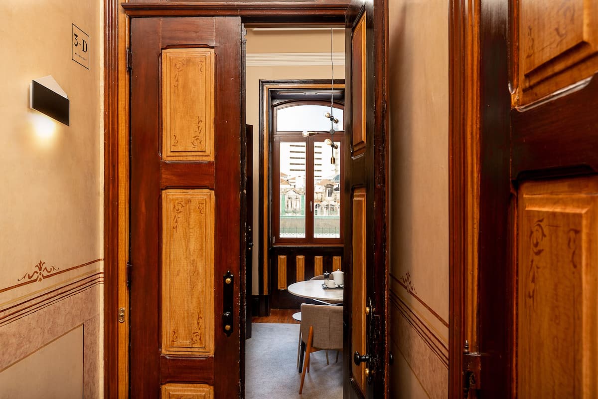 A narrow hallway with wooden doors and a small table with a chair, leading to a window with a view of buildings.