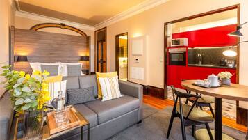 A modern living room with a grey sofa, a coffee table, and a dining table with chairs. There is a kitchen visible in the background with red cabinets.