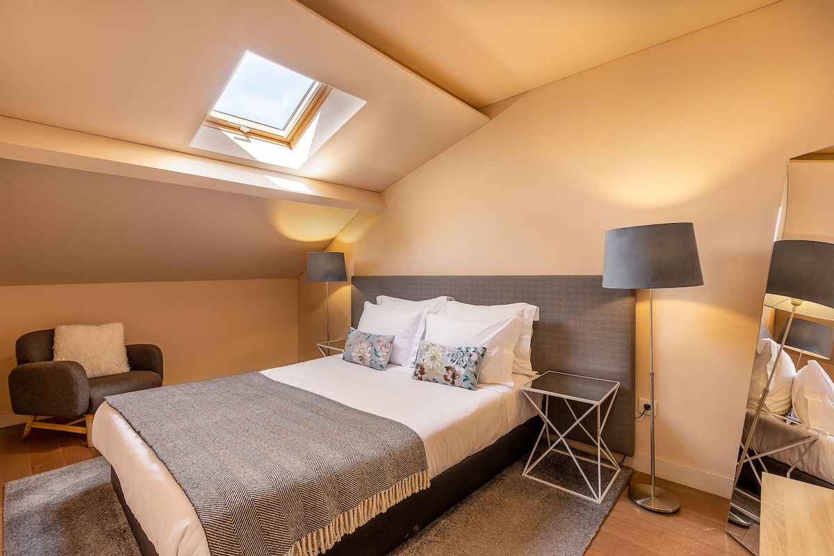 A modern hotel room with a skylight, a bed with white linens, a dark armchair, and a floor lamp.