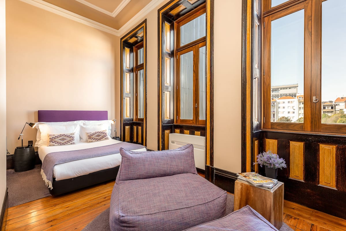 A hotel room with a large bed, a sofa, a small table with magazines, and large windows with wooden frames.