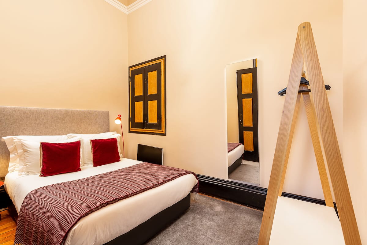 A hotel room with a bed, a mirror, a wooden ladder, and a door with a window.