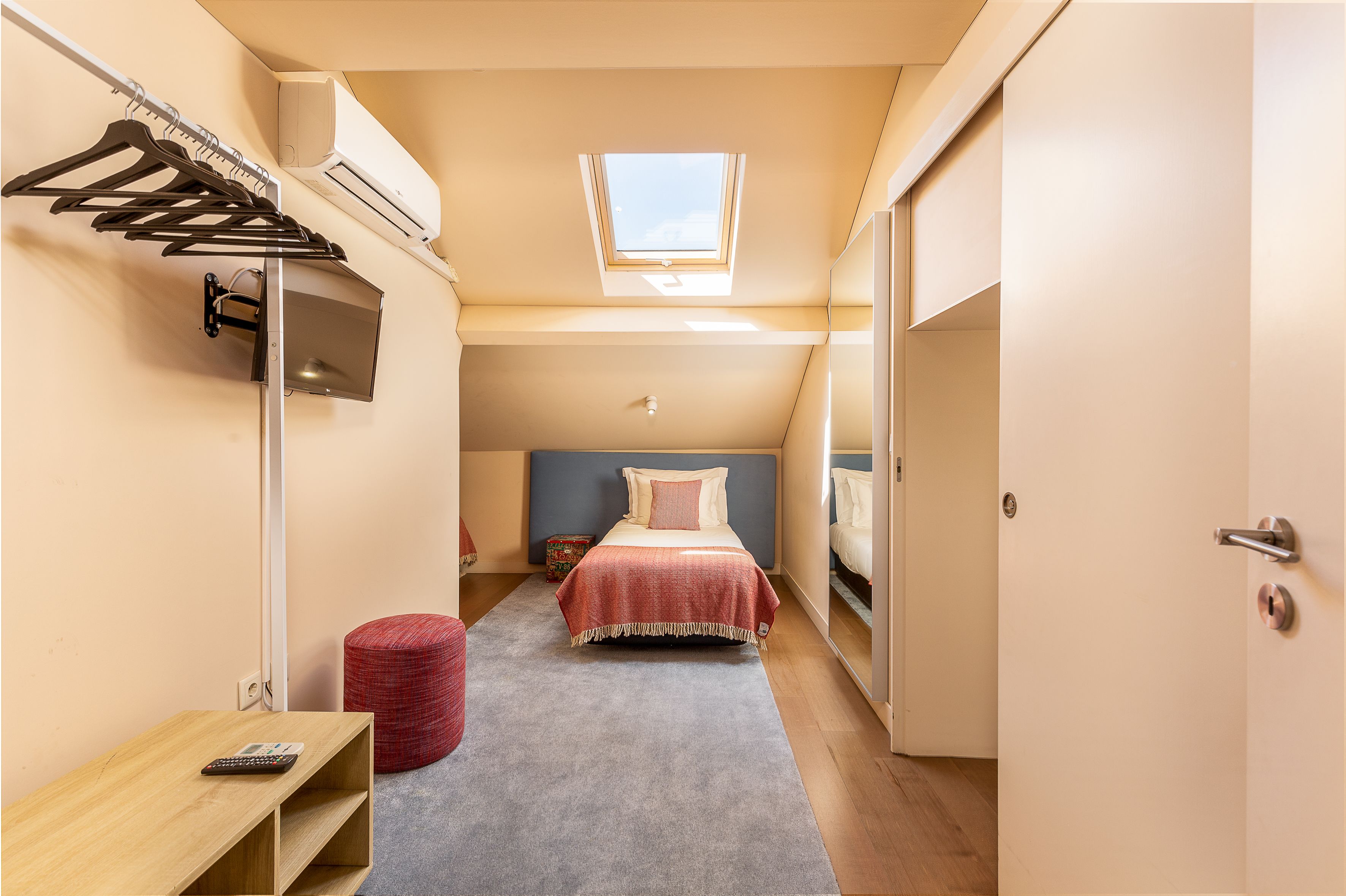 A bedroom with a bed, a red ottoman, a desk with a remote control, a wall-mounted TV, and an air conditioning unit.