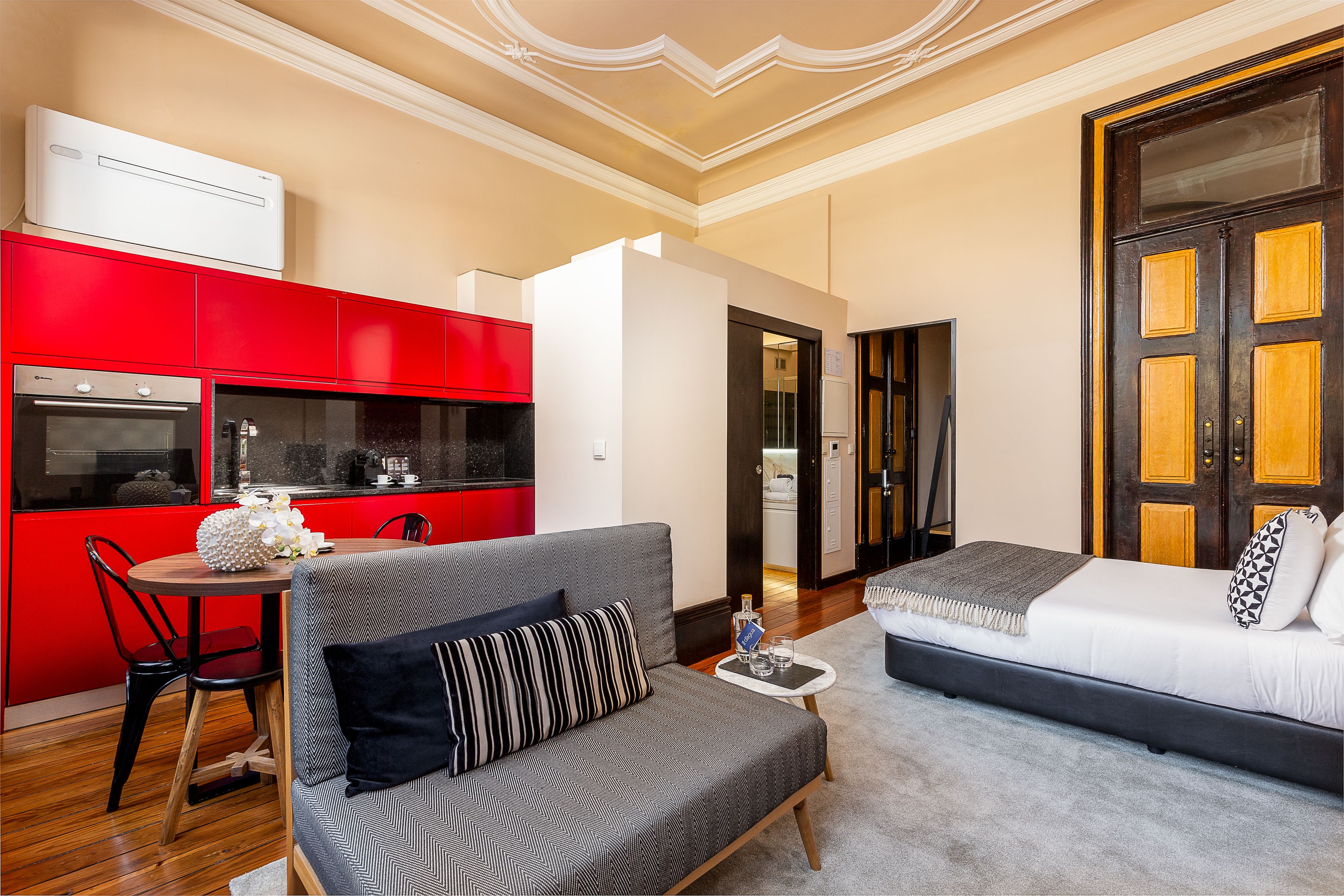 Een moderne woonkamer met een grijze bank, een eettafel met stoelen, en een rode keukenhoek.
