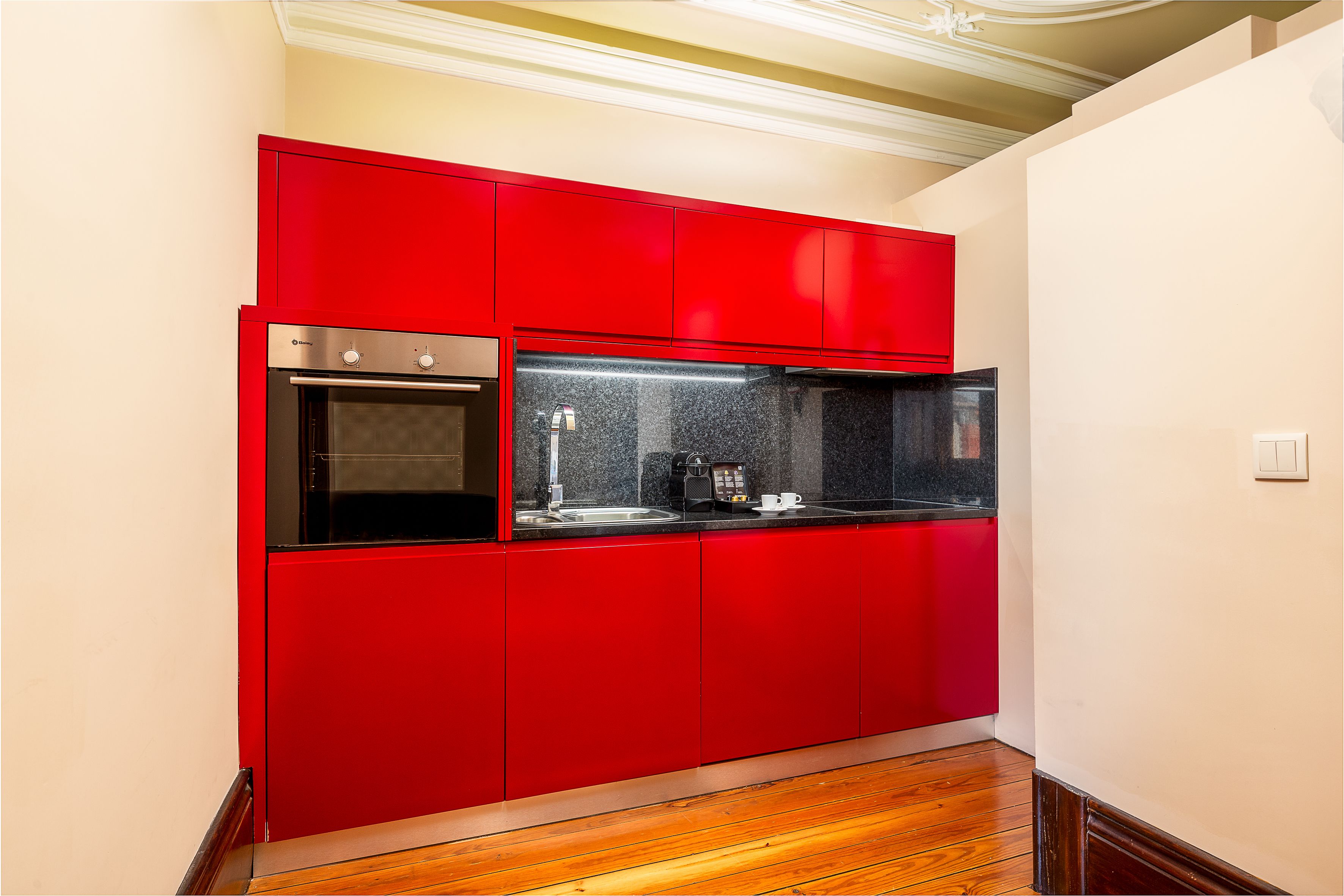 A compact kitchen with red cabinetry, a built-in oven, and a coffee maker on the countertop.