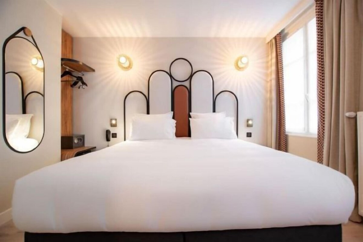 A large bed with a headboard, bedside tables, wall-mounted lights, and a window with curtains.