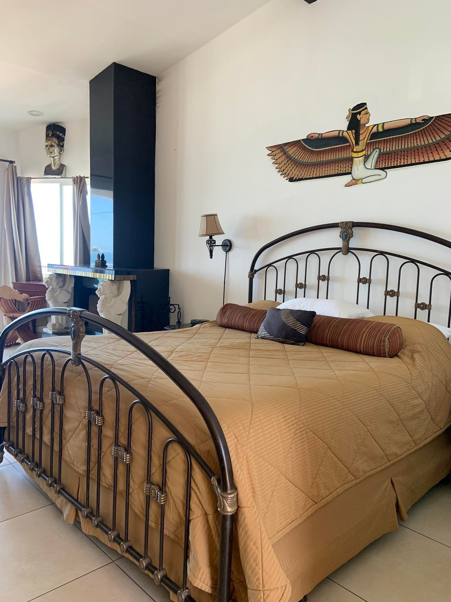 A bedroom with a metal bed frame, a large window with curtains, a wall-mounted lamp, and a decorative wall piece.
