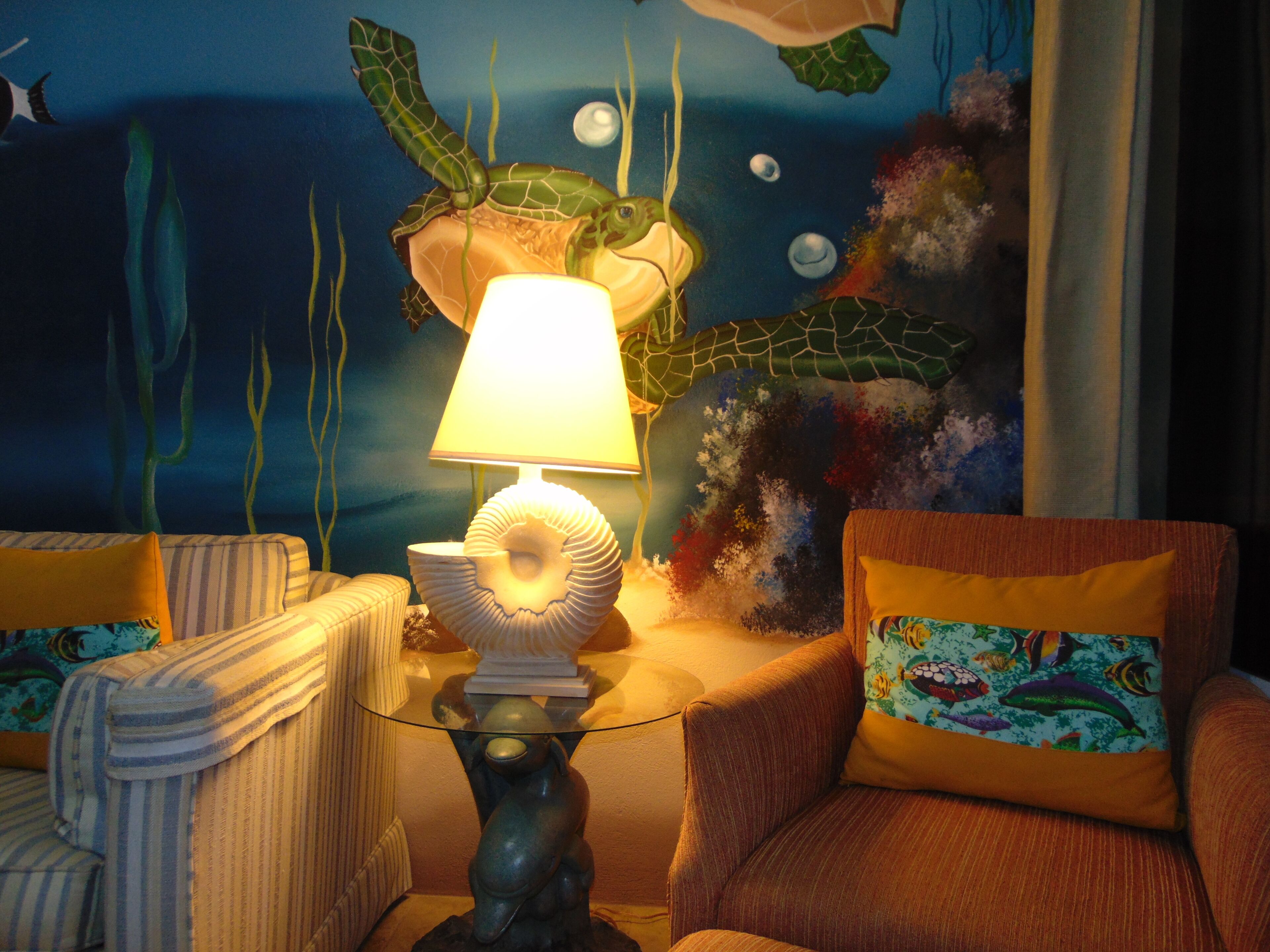 A room with a sea turtle mural, a lamp on a glass table, striped chairs, and colorful pillows.