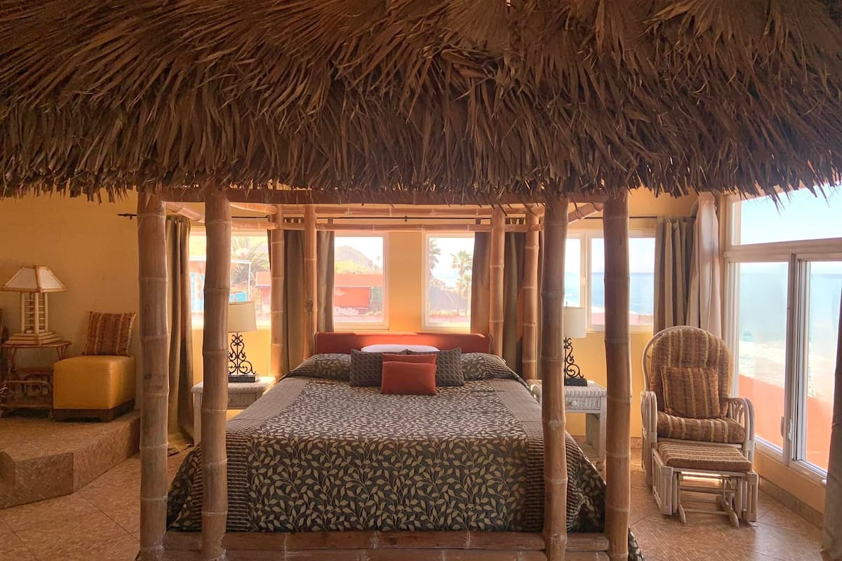 A room with a thatched roof canopy, a bed with patterned bedding, and a view of the sea.