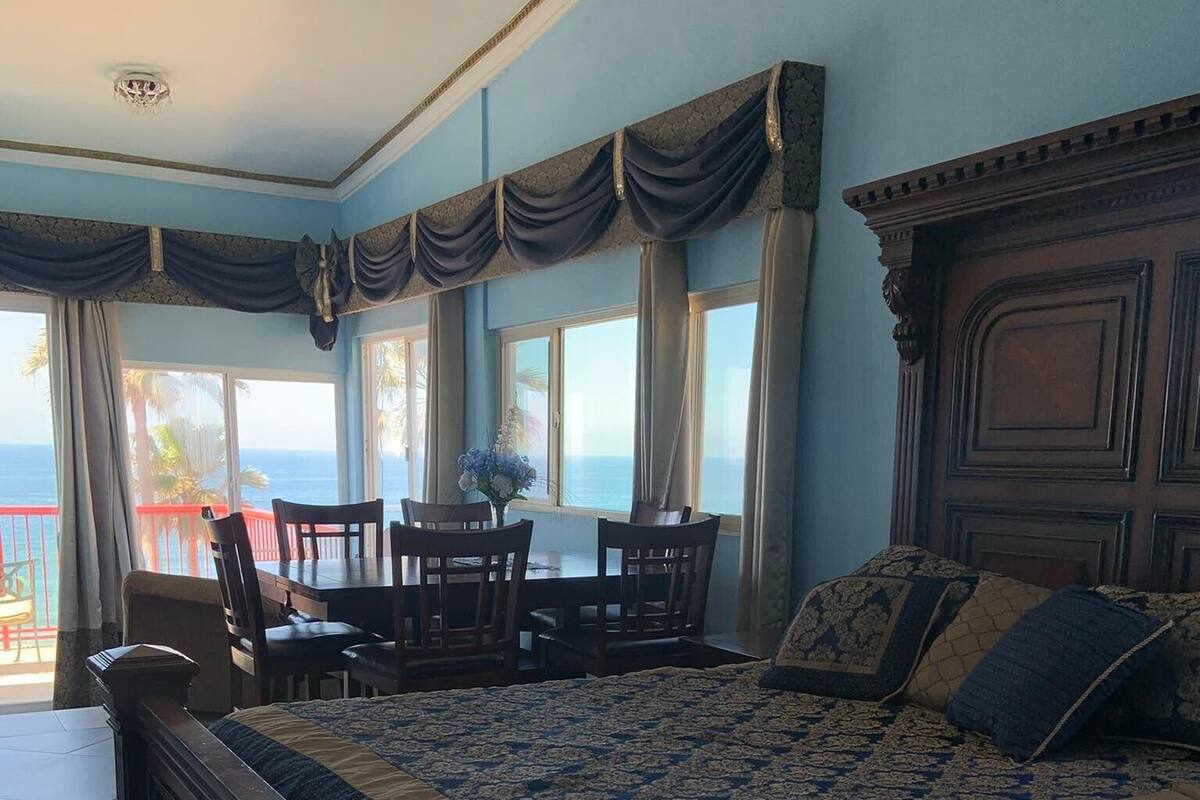 A bedroom with a large bed, a dining table, chairs, and a view of the ocean.