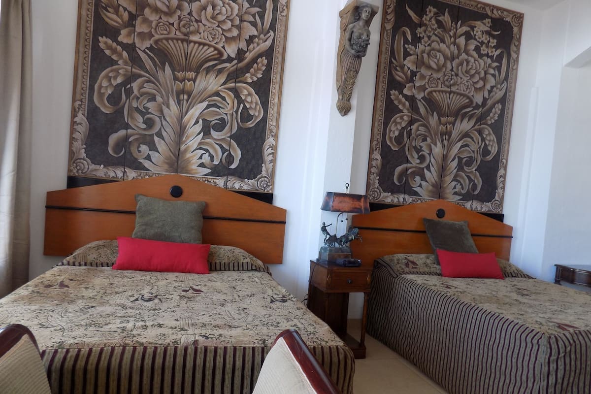 A hotel room with two beds, each with a wooden headboard, a nightstand with a lamp, and a large floral tapestry on the wall.