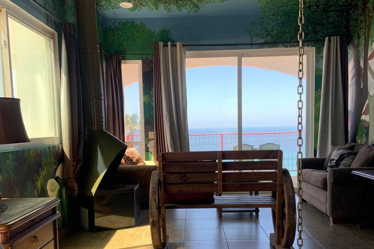 A room with a bed, a wooden bench, a sofa, and a view of the sea.