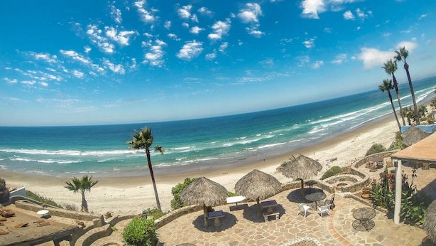 Vista de una playa con palmeras, sombrillas de paja y zonas de descanso de piedra.