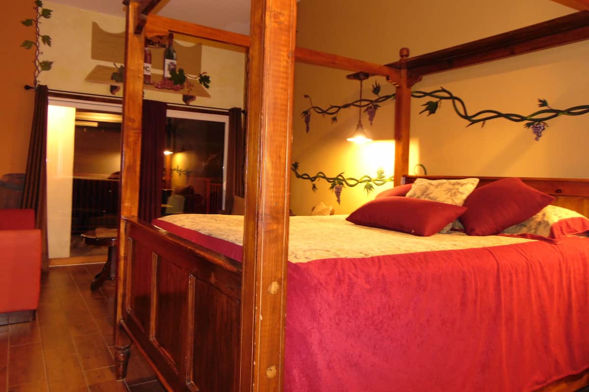 A four-poster bed with a red bedspread and two red pillows, a wooden ceiling with exposed beams, and a wall with grapevine decorations.