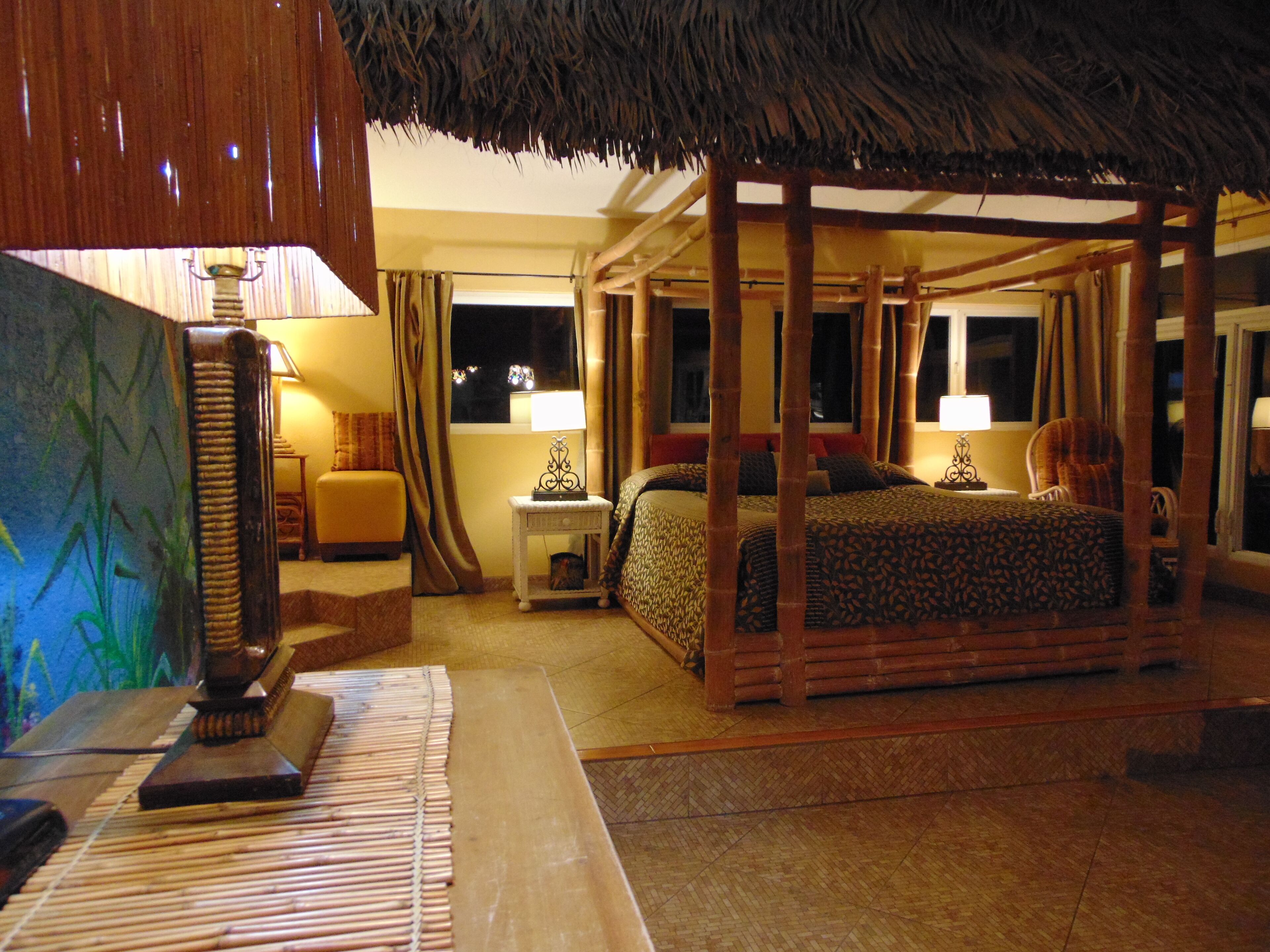A room with a four-poster bed, a nightstand with a lamp, a chair, and a large window with curtains.