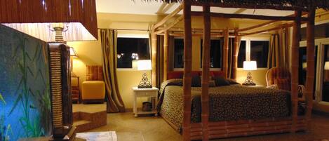A room with a four-poster bed, a nightstand with a lamp, a chair, and a large window with curtains.