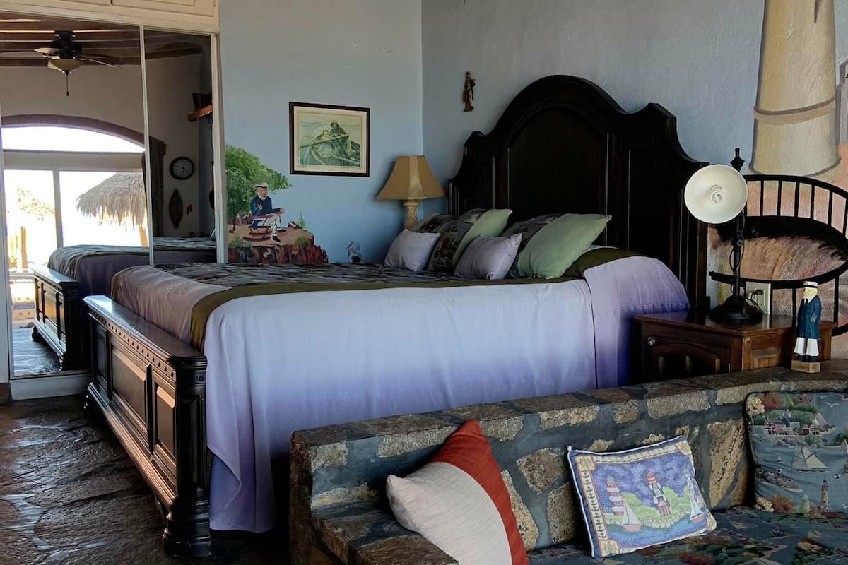 A bedroom with a bed, a nightstand, a lamp, a stone bench, and a wall with a lighthouse mural.