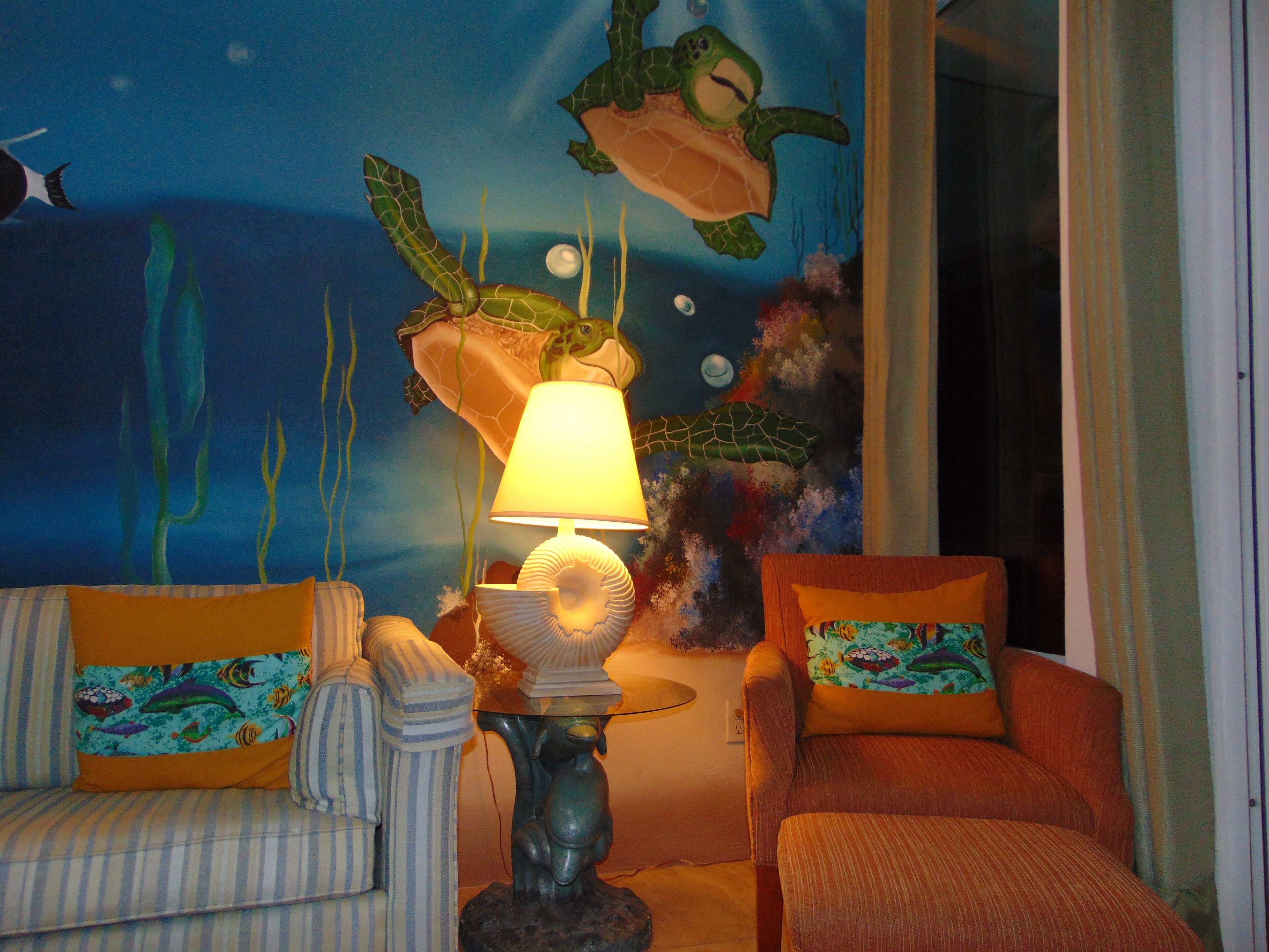 A room with a mural of an underwater scene, a lamp on a table, striped chairs, and a cushion with a fish design.