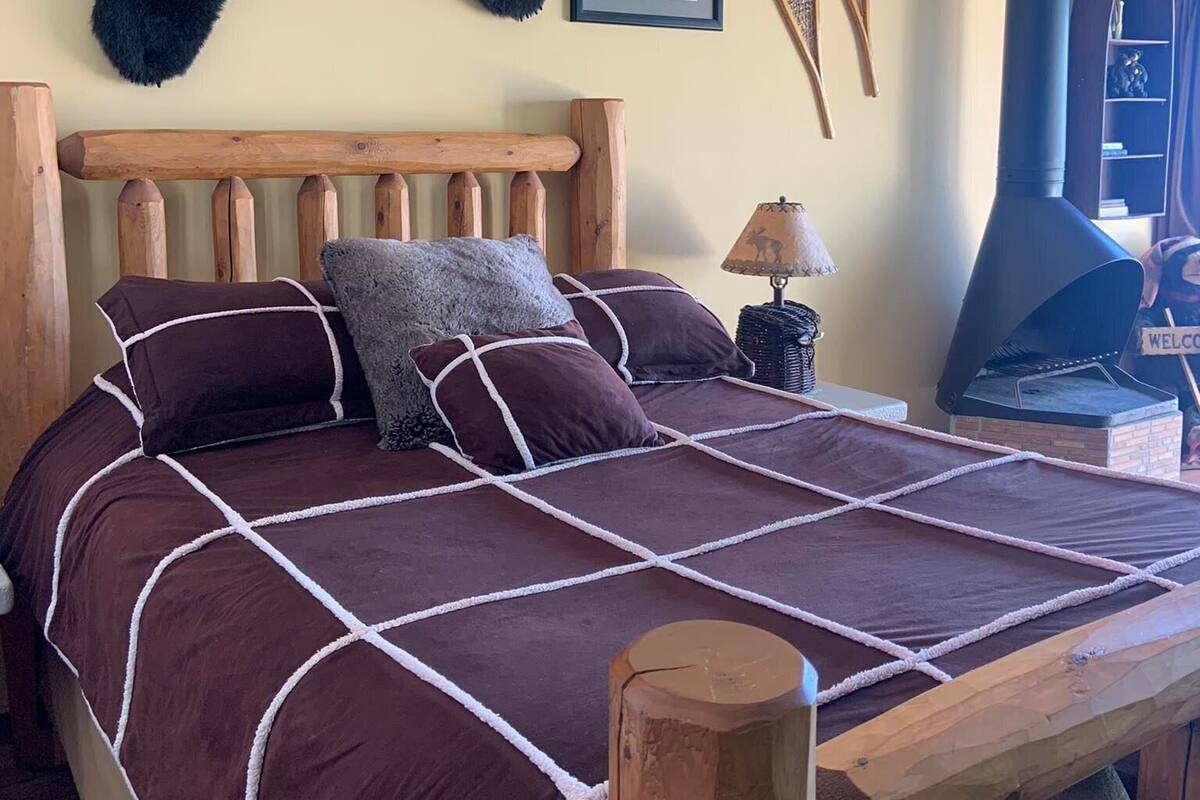 A bed with a wooden headboard, a large fur rug, a framed picture, a lamp, and a wood-burning stove.