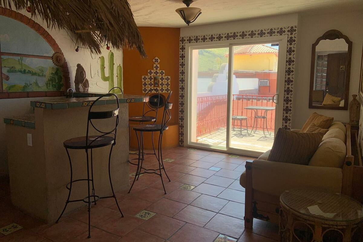A living area with a sofa, a small round table, and two bar stools. There is a large window with a view of a red fence and a balcony.