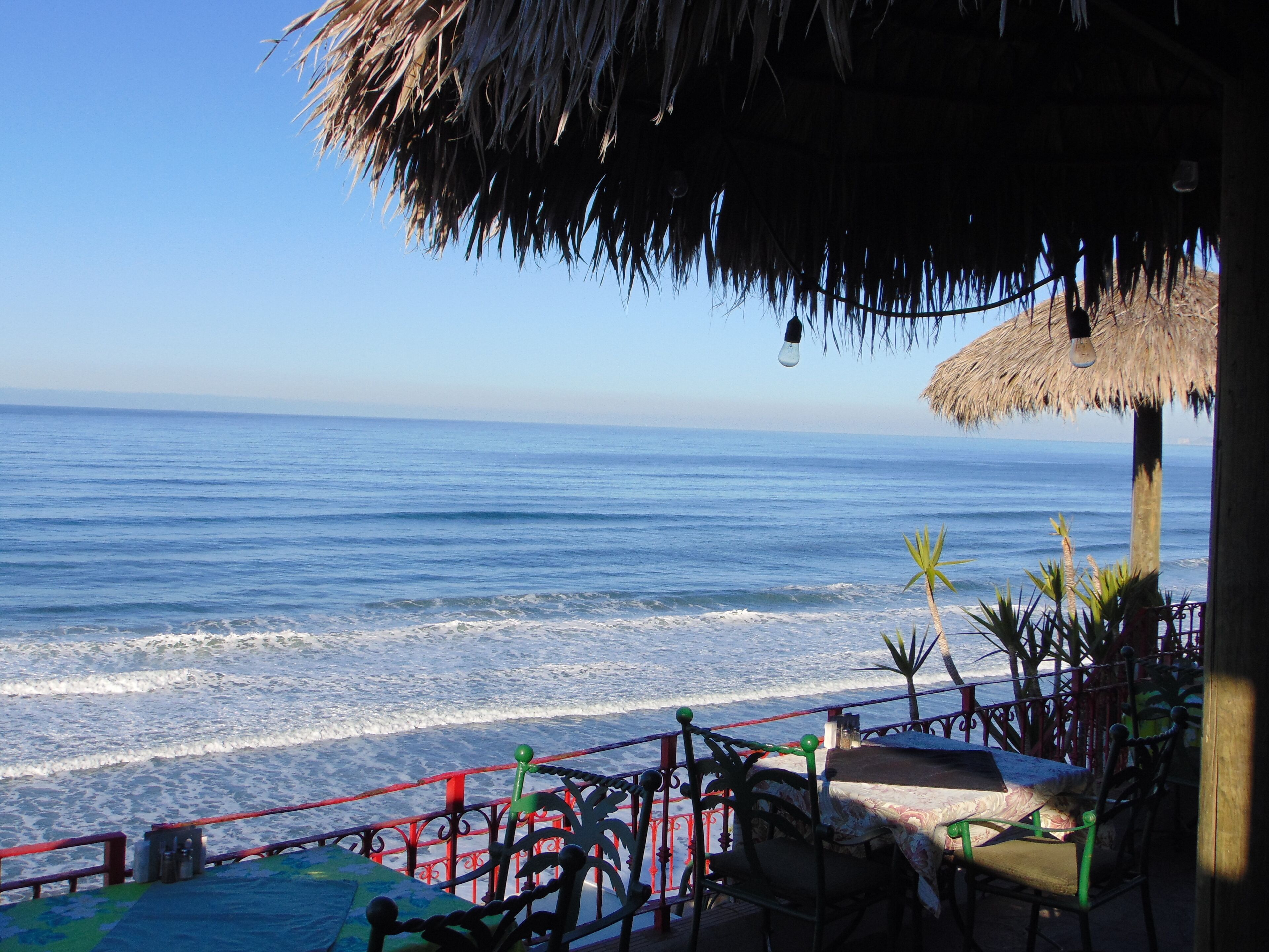 A beach view from a thatched-roof structure with a table and chairs, overlooking the ocean.
