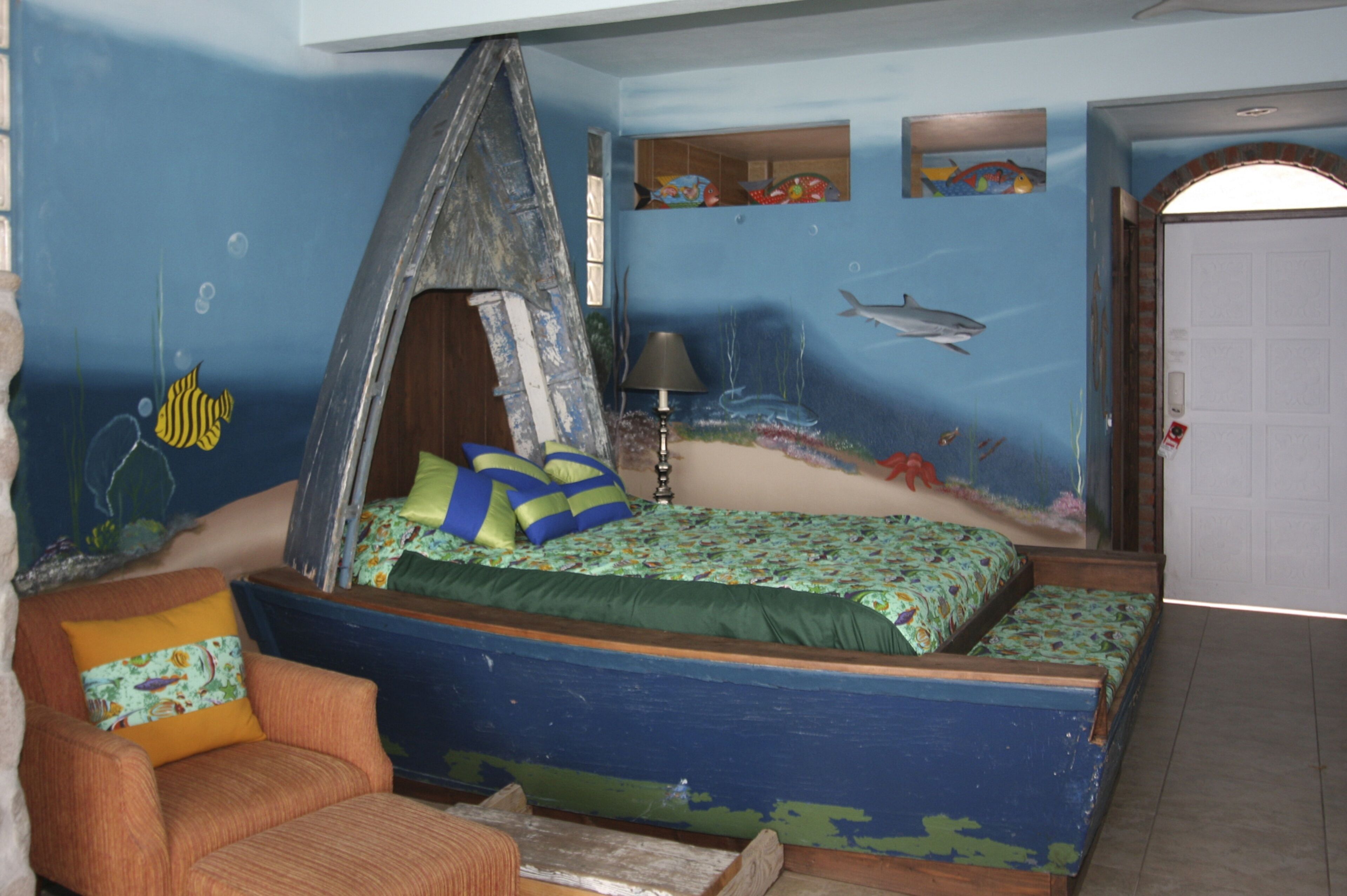 Quarto com cama em formato de barco, uma cadeira e um mural na parede retratando uma cena subaquática.
