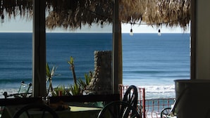 Vista desde un área techada junto a la playa, con techo de paja, un pilar de piedra y barandilla con vista al océano.