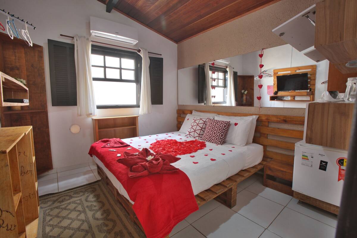 Un dormitorio con cama, cabecera de madera, ventana con cortinas, espejo, televisor y refrigerador.
