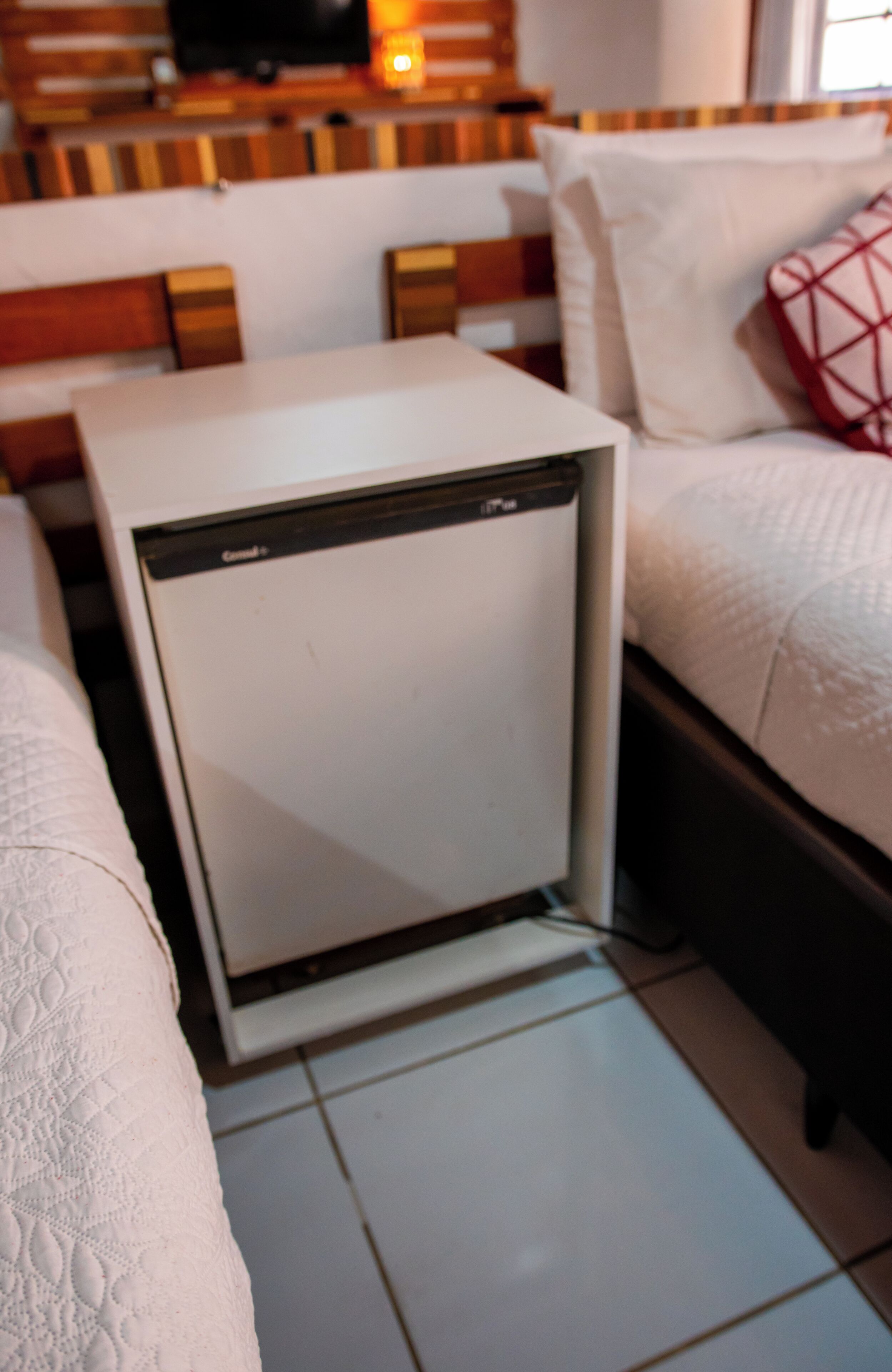A compact room with a white refrigerator, a bed with white bedding, and a wooden headboard.