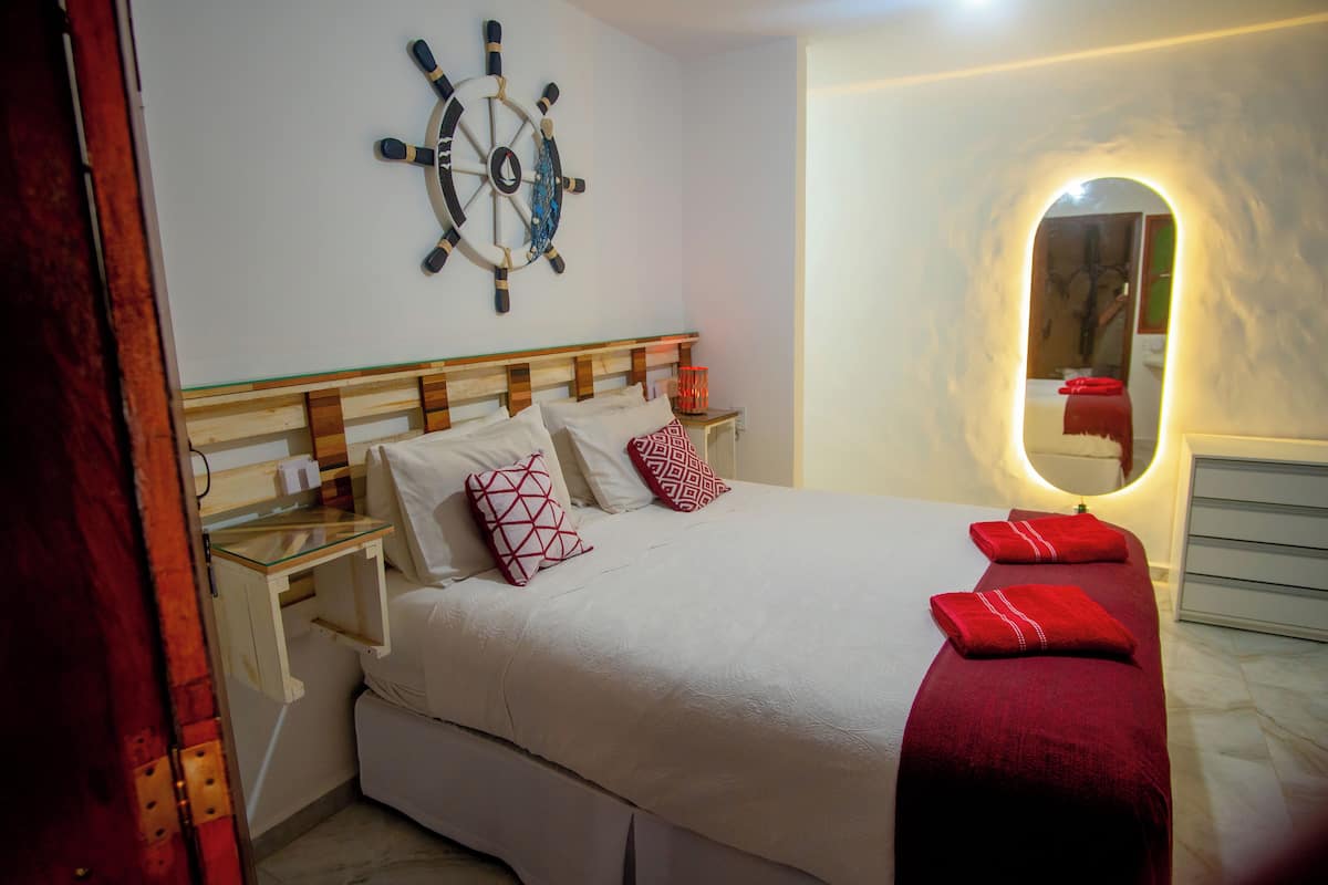 Una cama bien hecha con cabecera de madera, dos mesitas de noche con lámparas, un adorno de timón de barco en la pared y un espejo con marco circular.