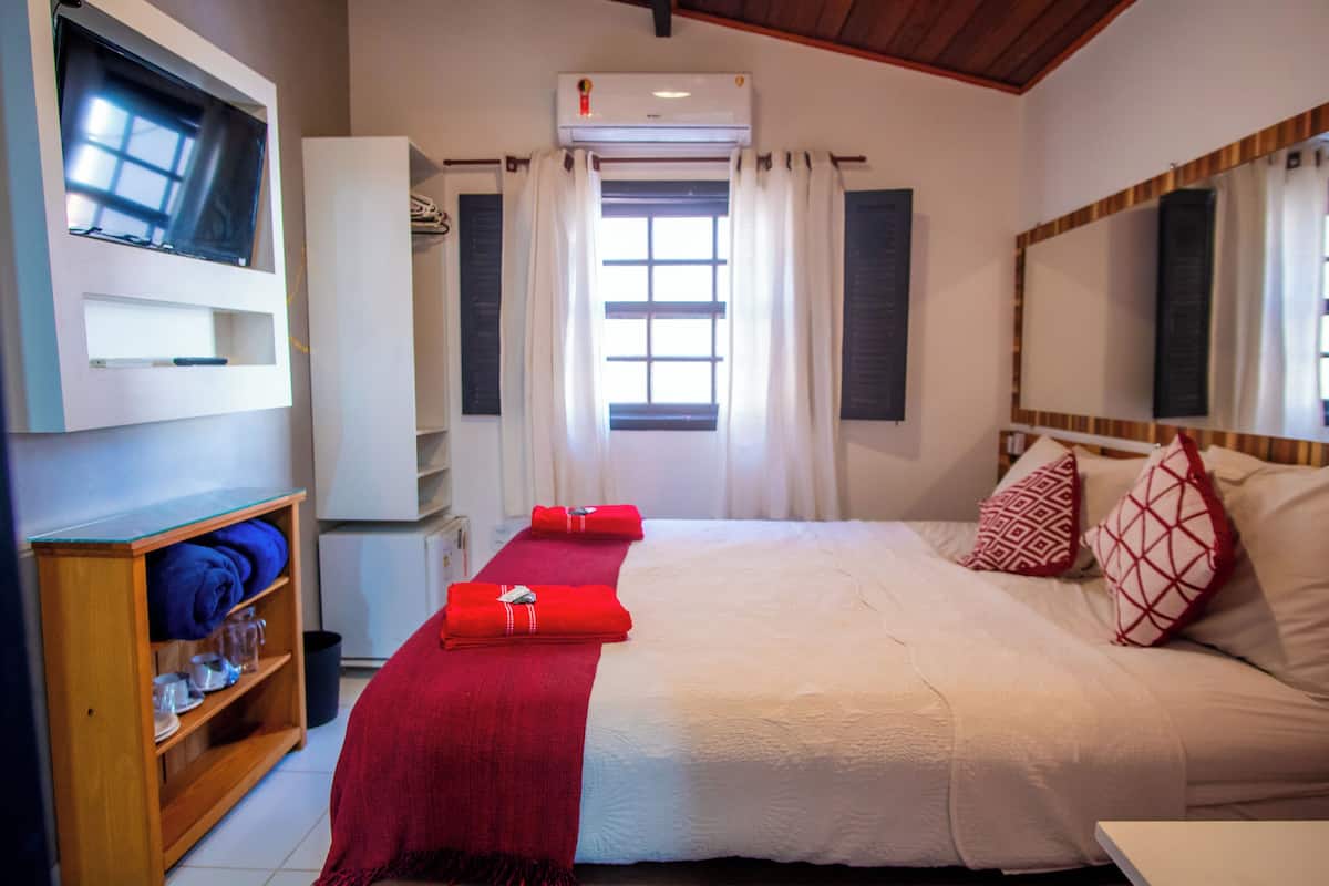 Una habitación de hotel con cama, televisor montado en la pared, armario y ventana con cortinas.