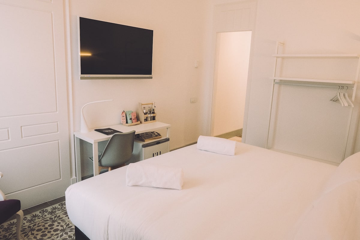 A hotel room with a bed, a desk with a chair, a flat-screen TV mounted on the wall, and a wardrobe with hanging clothes.