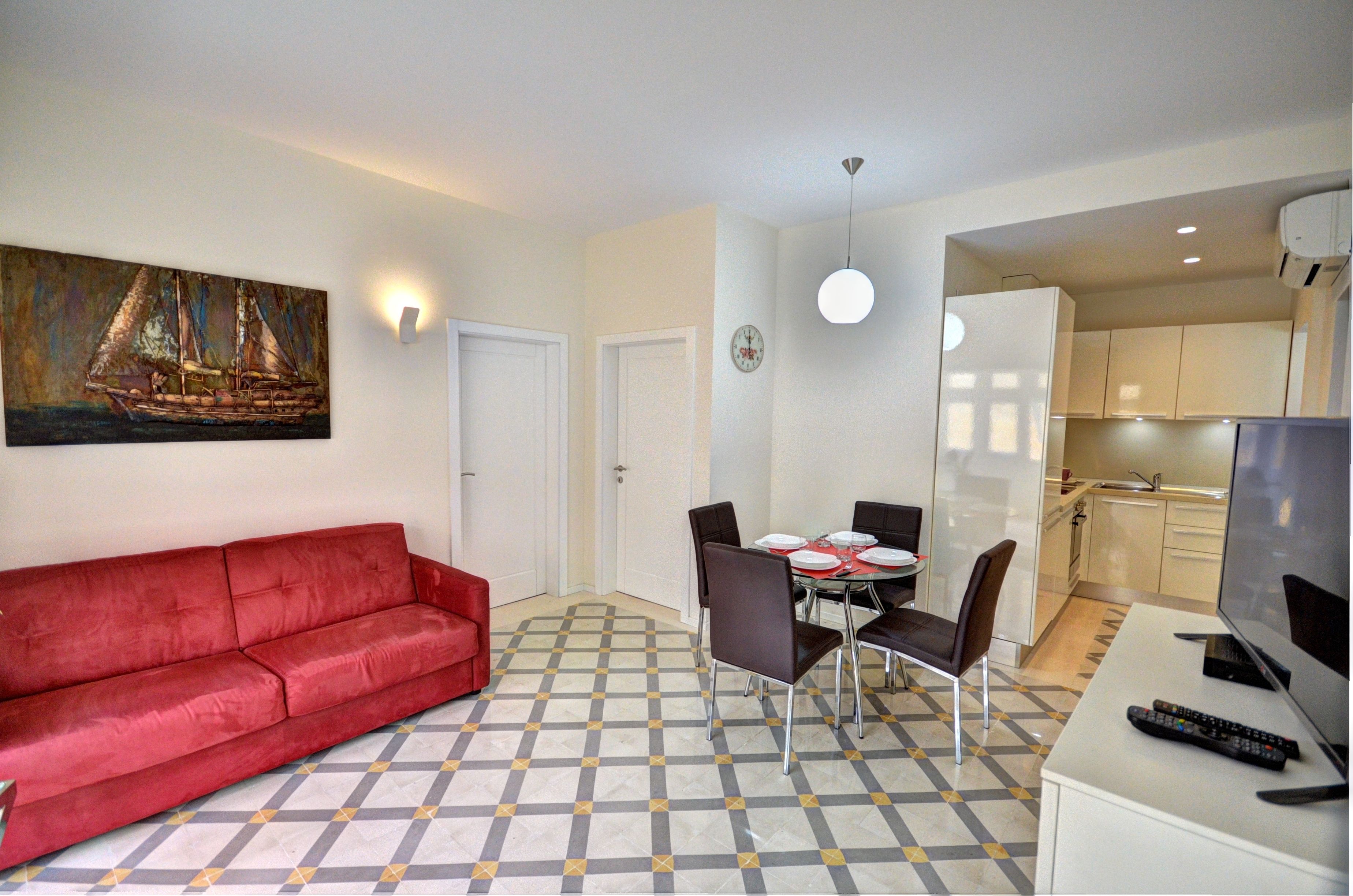 A living room with a red sofa, a dining table with chairs, a TV, and a kitchen area in the background.