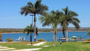 A lakeside view with palm trees, a dock, and a boat. - Marina Flat & Náutica Apartamento - Férias em Caldas Novas (Caldas Novas)