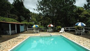 A rectangular swimming pool with stone tiles, surrounded by white plastic chairs and tables under umbrellas. - Pousada & Camping Serras Verdes (Valenca)