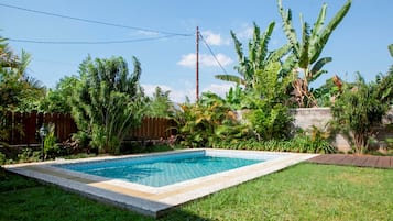 Piscina rectangular con borde de azulejos, rodeada de césped y diversas plantas tropicales.