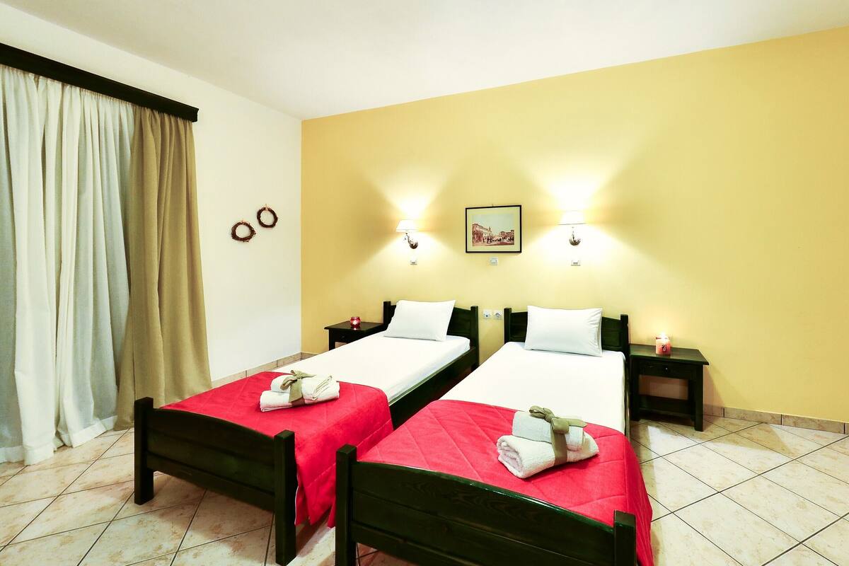 A hotel room with two single beds, each with a red bedspread and white pillows, a nightstand with a lamp, and a picture on the wall.
