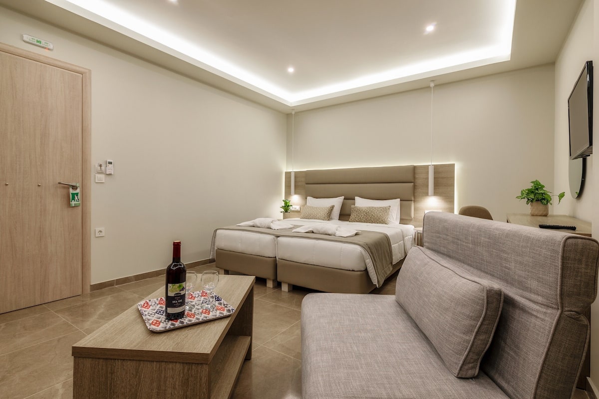 A modern hotel room with a large bed, a sofa, a coffee table with a bottle of wine, and a small table with a plant.