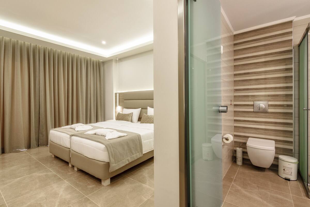 A modern hotel room with a large bed, a separate sofa, a glass shower enclosure, and a bathroom with a striped tile backsplash.