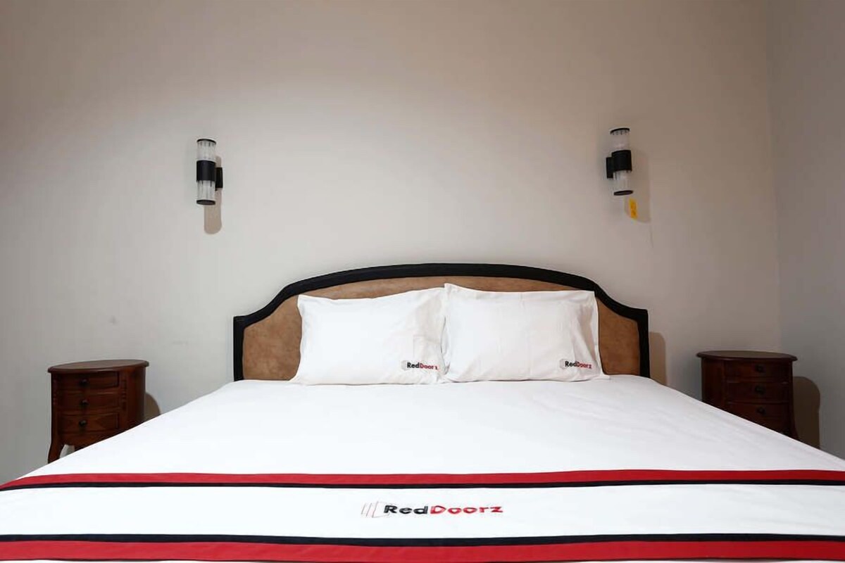 Een bed met een witte matras en een rood-zwart gestreept dekbed, twee nachtkastjes en een wandlamp.