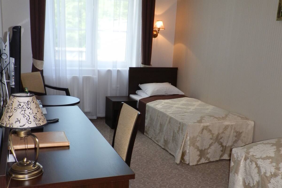 Une chambre d’hôtel avec un bureau en bois, deux lits simples, une chaise, une lampe et une fenêtre avec des rideaux.