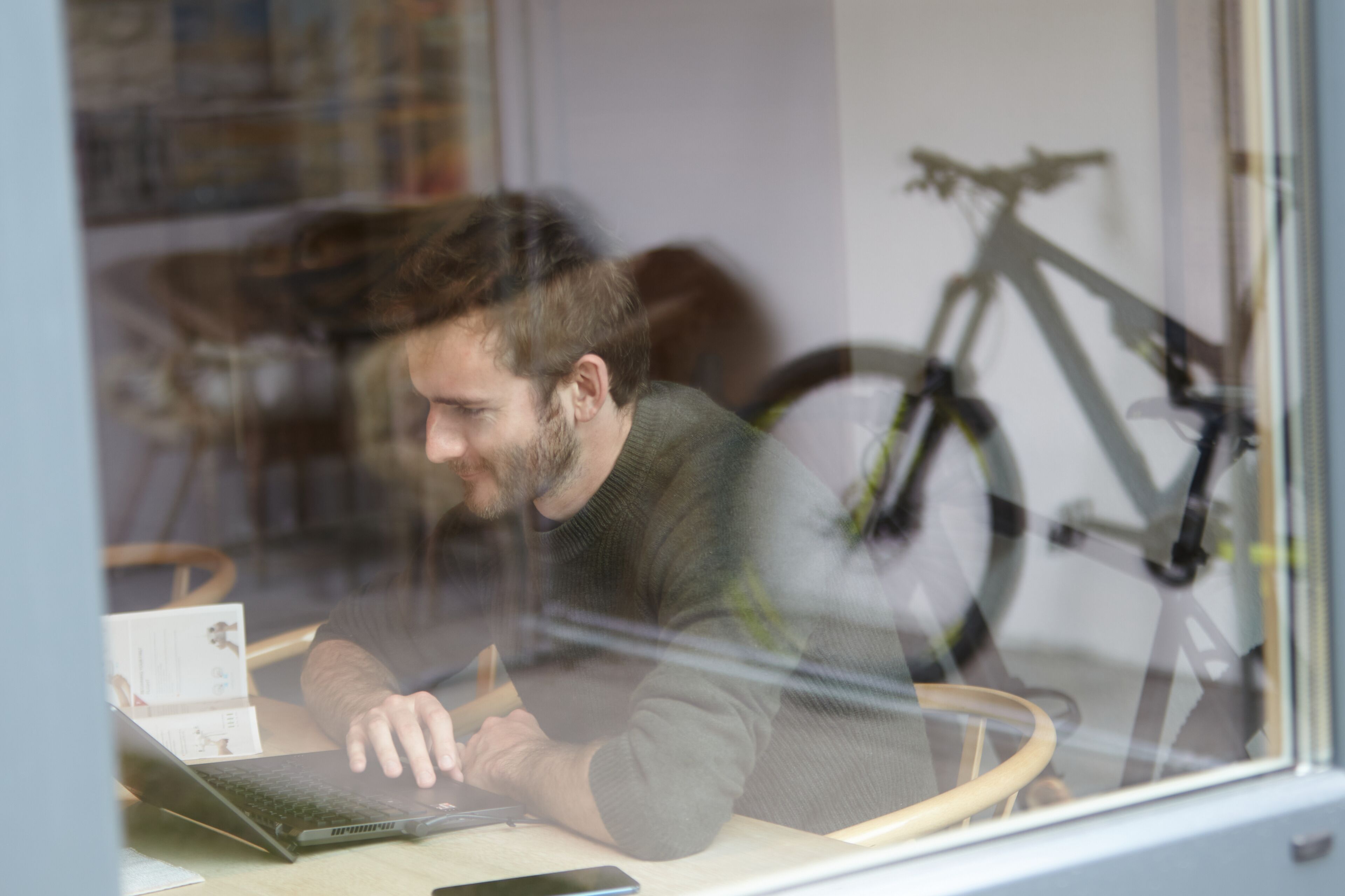 A person working at a desk with a laptop, a smartphone, and some papers. A bicycle is visible in the background.