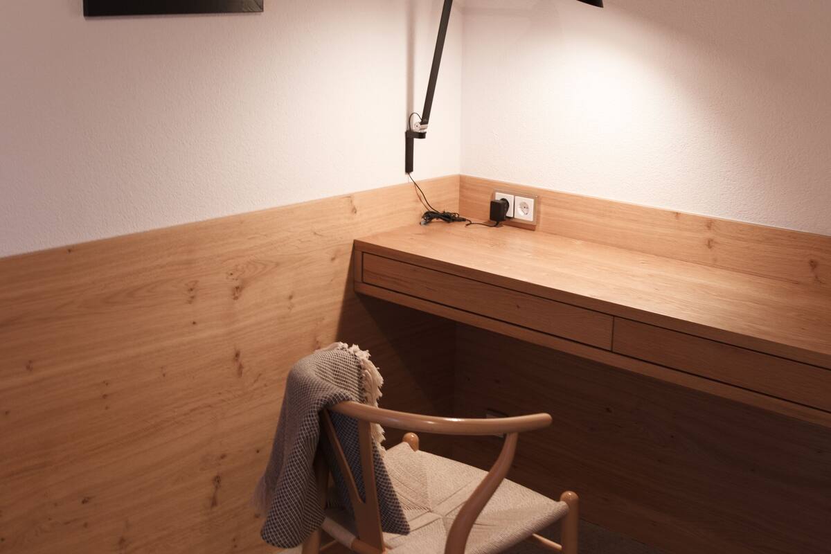 A corner workspace with a wooden desk, a chair, a framed picture, and a desk lamp.