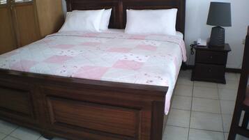 Una cama de madera con colcha, una mesita de noche con lámpara y una silla.